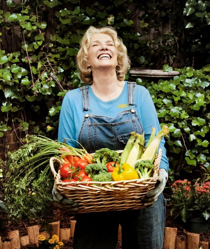 blonde Frau mit Gemüsekorb in den Händen vor Gebüsch
