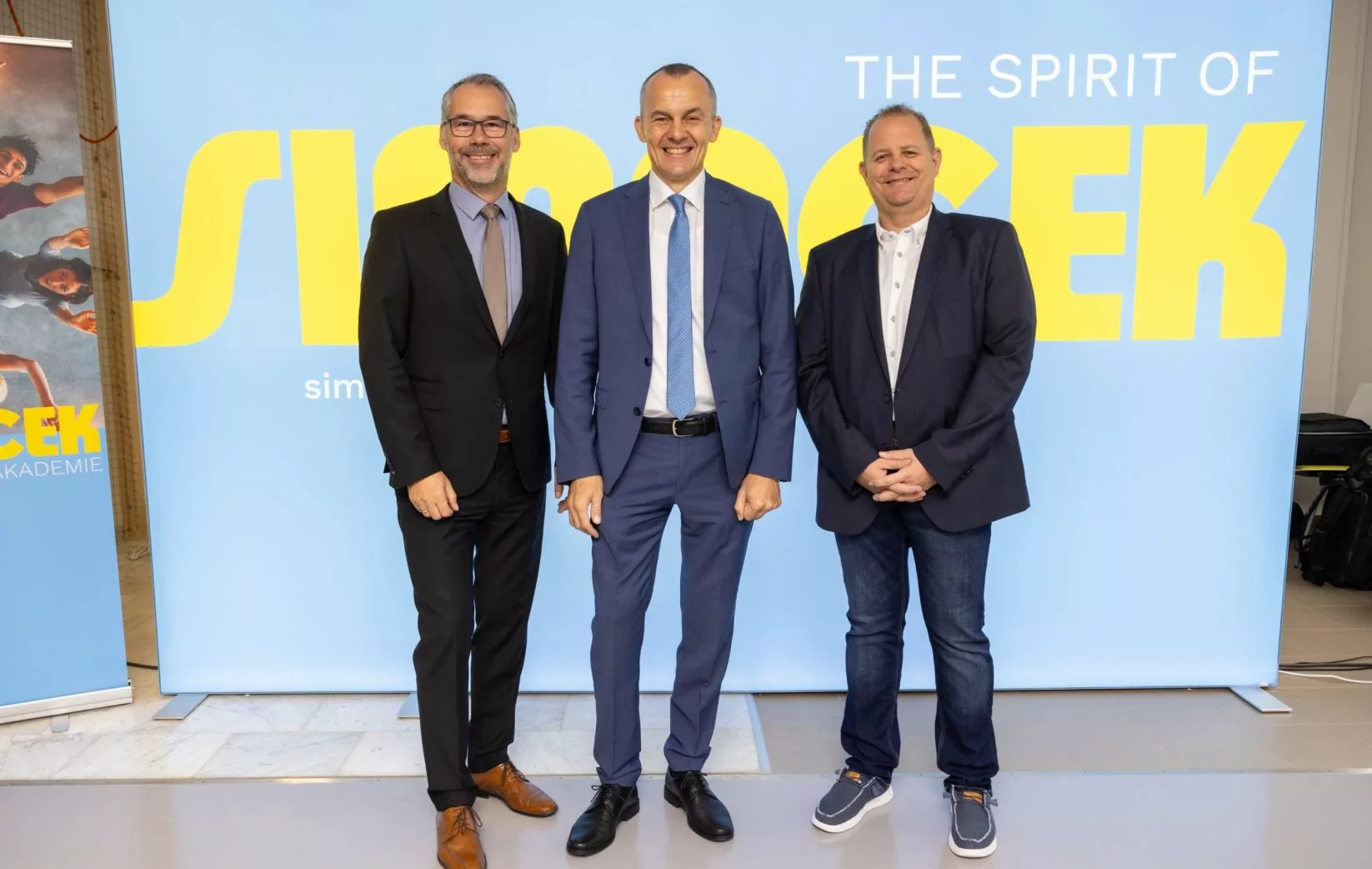 Drei Herren stehen vor "The Spirit of Simacek"-Wand